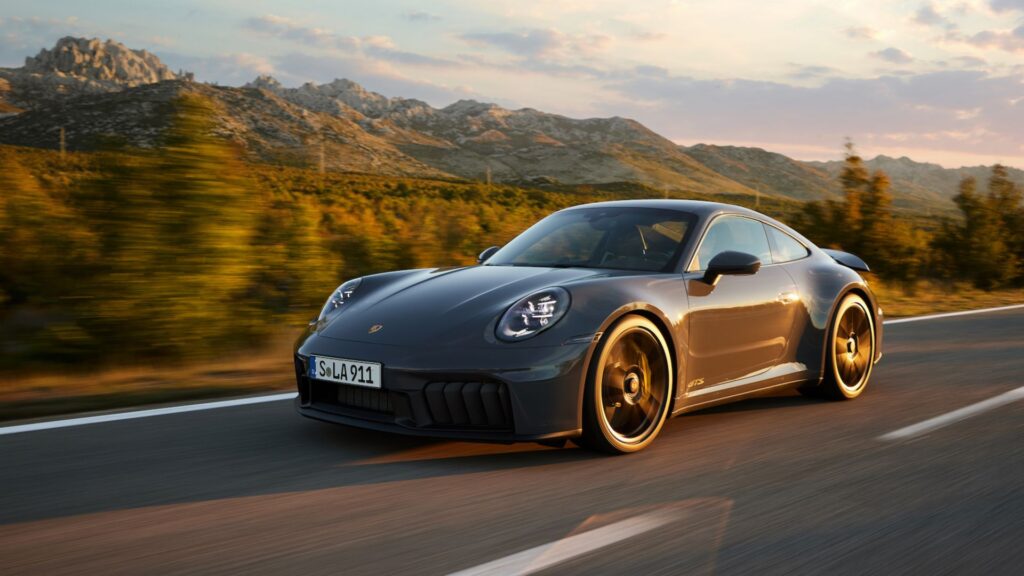 A grey Porsche 911 GTS driving on a winding mountain road at sunset, with blurred golden foliage in the background.