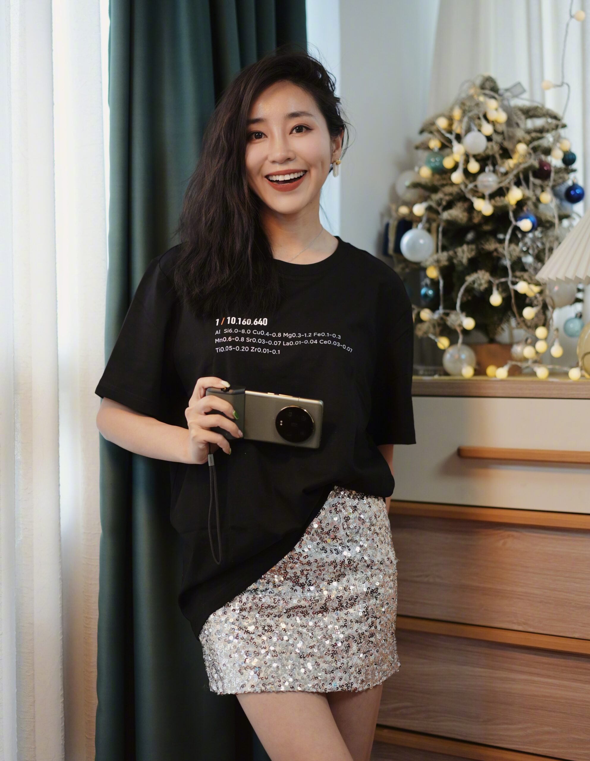 A cheerful woman holding a Xiaomi 15 Ultra, wearing a black t-shirt with scientific text and a glittering sequin skirt.