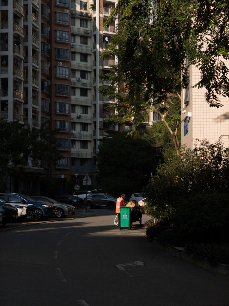 A quiet residential street surrounded by buildings, trees, and a person tending to trash bins in the afternoon light.