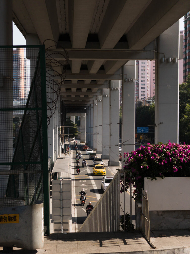 A street view under an elevated bridge, featuring parked taxis and colorful flower arrangements.