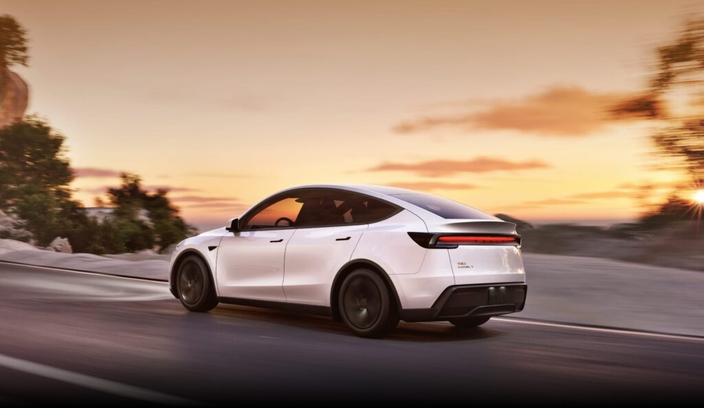 Rear view of the Tesla Model Y facelift in white, driving on a scenic road at sunset, highlighting its sleek design and new rear light-bar.