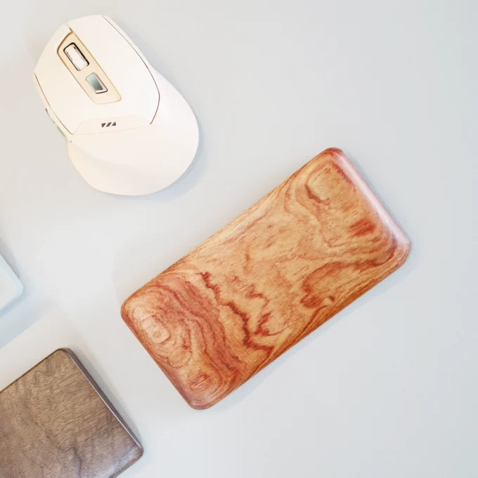 The Artisan Wood Ergonomic Mouse Wrist Rest, displaying its detailed wood grain pattern, lying beside a white mouse and a keyboard edge visible in the background.