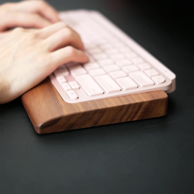 Side angle of the pink keyboard positioned on the Walnut Wood Wrist Rest Tray, emphasizing the tray’s curved profile and rich walnut texture, with hands in the background for scale.