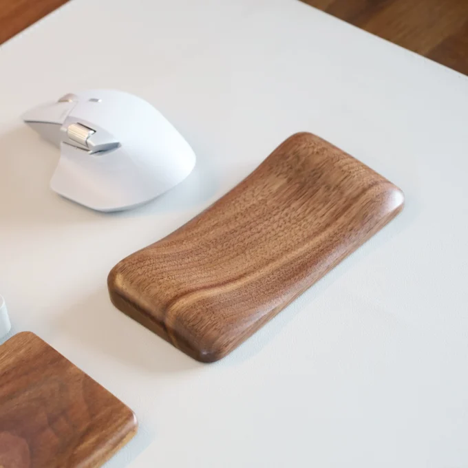 Two Artisan Wood Ergonomic Mouse Wrist Rests in different shades lying on a white surface near a white computer mouse, showing the variety of wood finishes.