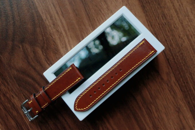 The brown leather Apple Watch band laid over a phone, emphasizing its lustrous finish and bold contrast stitching.