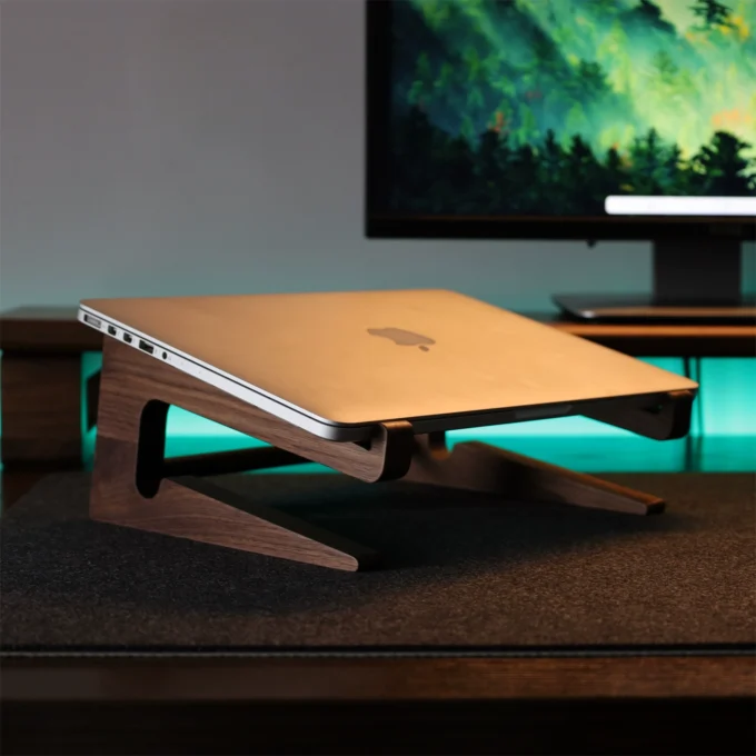 A walnut stand propping a MacBook at an ergonomic angle, with a forest-themed monitor backdrop.