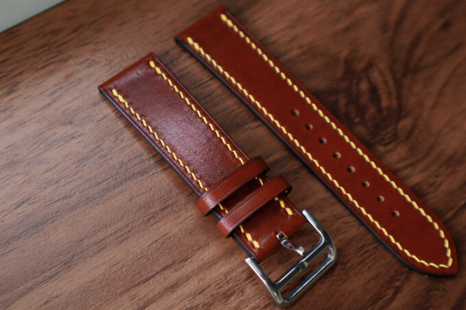 A brown Belgian Masure Shoulder Leather Apple Watch band with contrasting yellow stitching and a silver buckle, shown on a wooden tabletop.