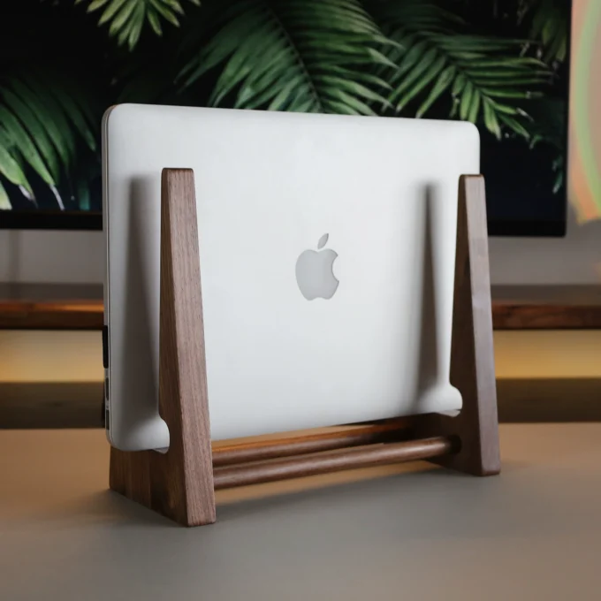 A closed MacBook stored vertically in a walnut wood stand on a desk, palm leaf décor in the background.