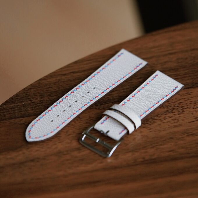 A white French leather Apple Watch band and its shorter buckle piece lying flat on a wooden table.