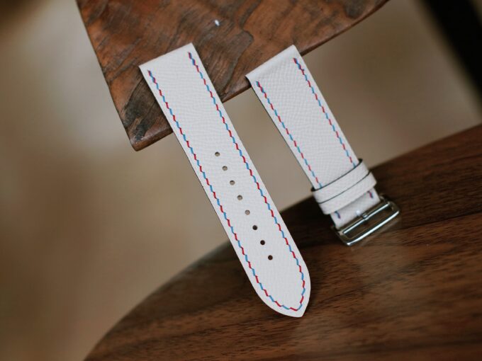 The band’s buckle side and longer strap displayed upright, revealing precise stitching along the white leather.