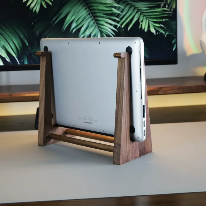 Close-up side view of a closed MacBook held upright by a walnut stand, showing the stand’s smooth wood grain.