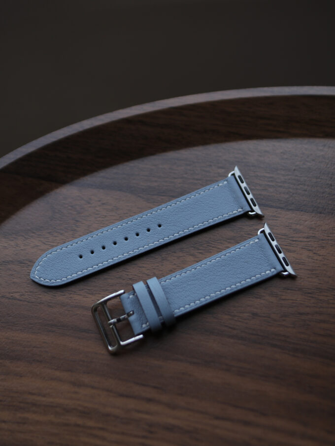 A blue Italian Pomari leather Apple Watch band and its buckle piece lying flat on a wooden table.