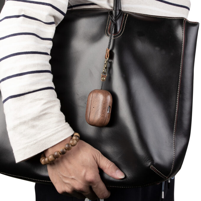 A walnut wood AirPods case hanging securely on a black leather handbag using the natural leather strap, showing its portability and minimalist design.