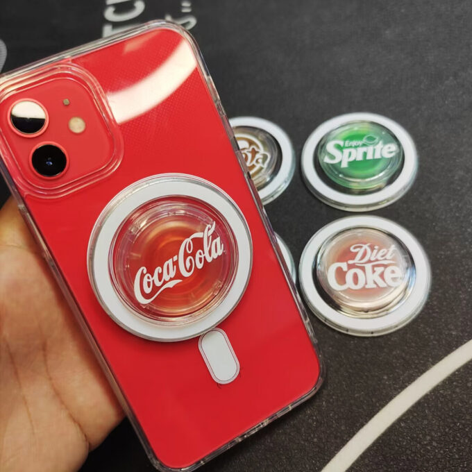 Another red iPhone showcasing a Coca-Cola MagSafe grip, with additional beverage-themed grips (Diet Coke, Sprite) arranged around it.