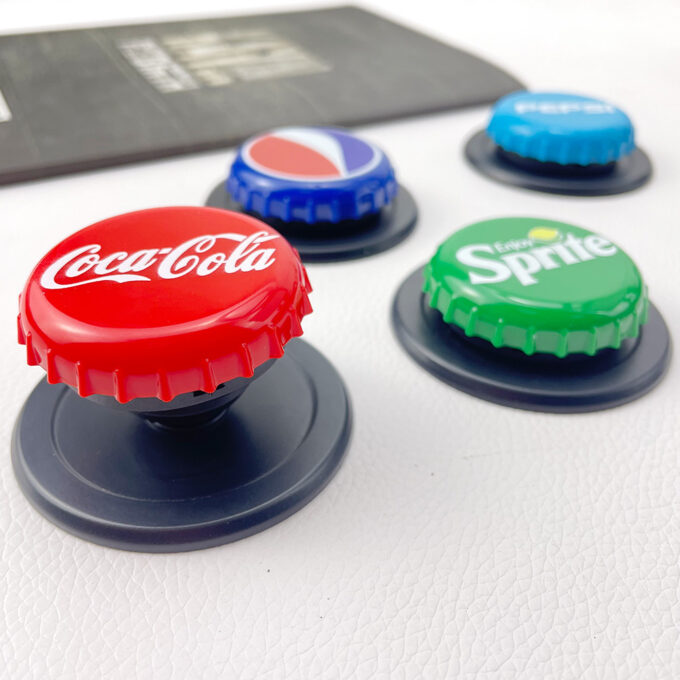 A gray iPhone with a Coca-Cola bottle cap grip attached, surrounded by additional caps in Sprite and Fanta designs.