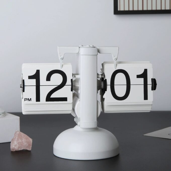 A classic white mechanical flip desk clock showing “12:01 PM,” set against a dark desk with a contemporary aesthetic.