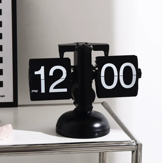 A sleek black mechanical flip desk clock displaying “12:00 PM,” placed on a modern metal table for a bold and minimalist look.