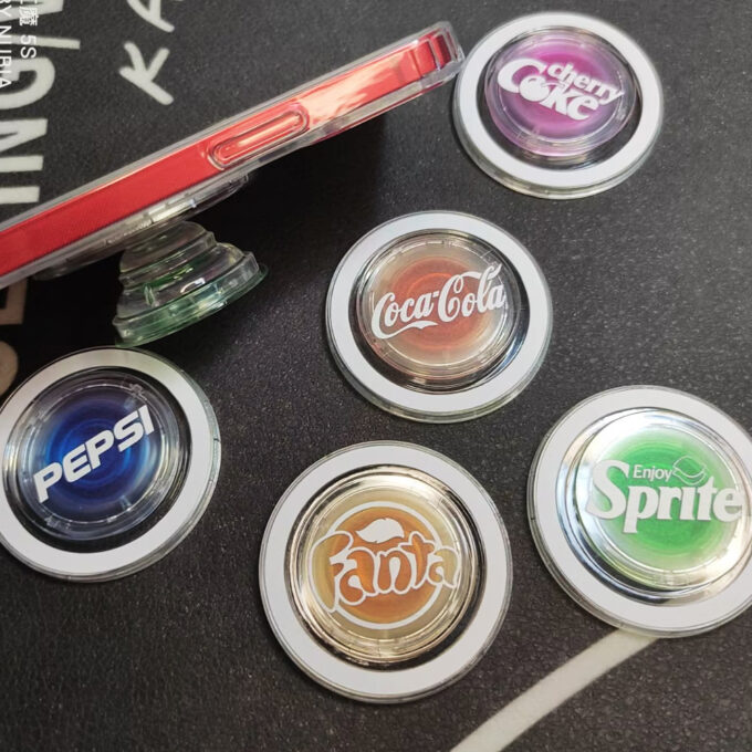 Multiple circular MagSafe grips laid out, featuring Coke, Fanta, Pepsi, and other printed designs, with the edge of a red phone in the background.