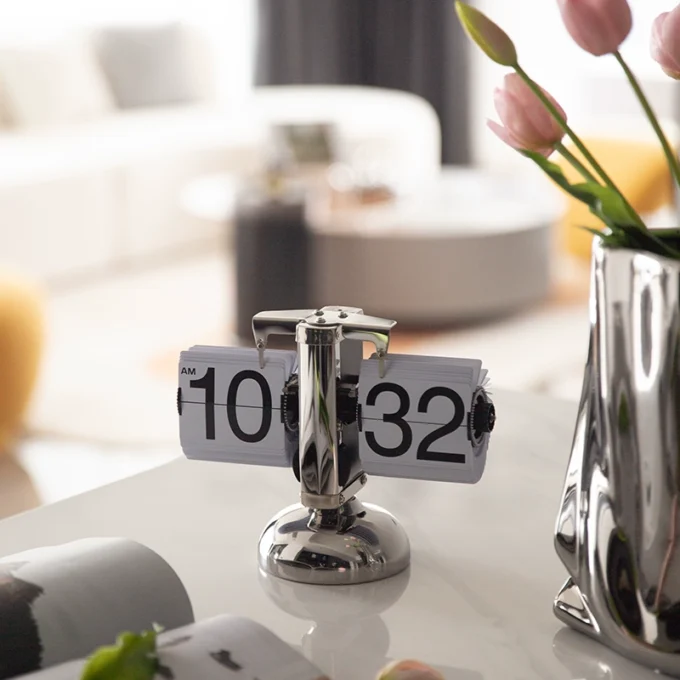 A mechanical flip clock with a polished silver finish, positioned in a bright and modern home setting.
