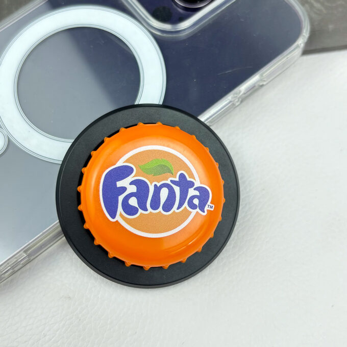 An orange Fanta bottle cap grip on a black stand next to a clear MagSafe phone case, highlighting the playful design.