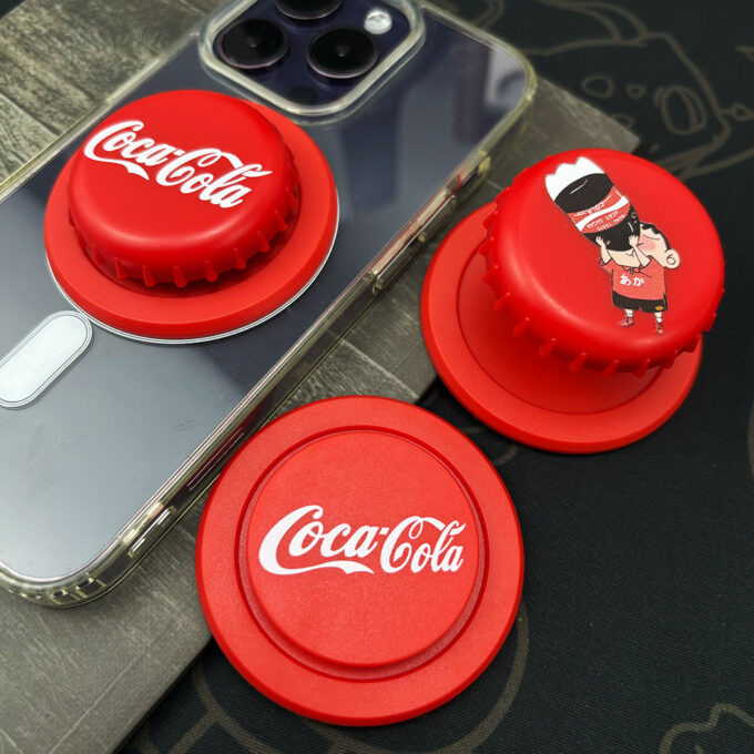 Three Coca-Cola bottle cap grips snapped onto a silver phone, with a separate red cap placed nearby, demonstrating their foldable functionality.