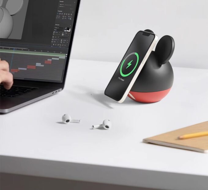 The mouse-eared charger on a desk next to a laptop, holding an iPhone in portrait mode while charging.