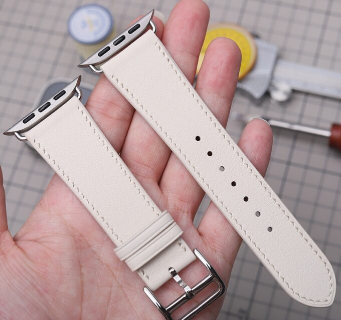 A close-up of a white French Swift Leather Apple Watch band, showing the stainless steel buckle and adapters in someone’s hand.