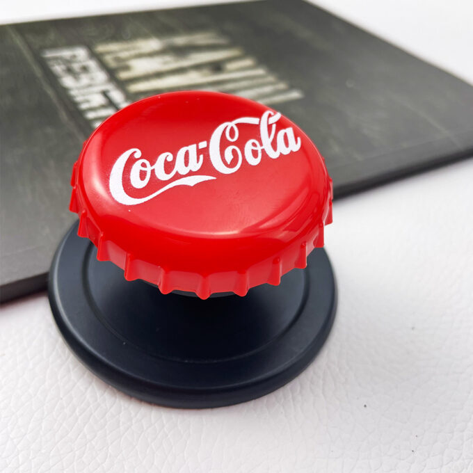 A red Coca-Cola bottle cap grip on its extended stand, set against a white backdrop with other brand-themed caps nearby.