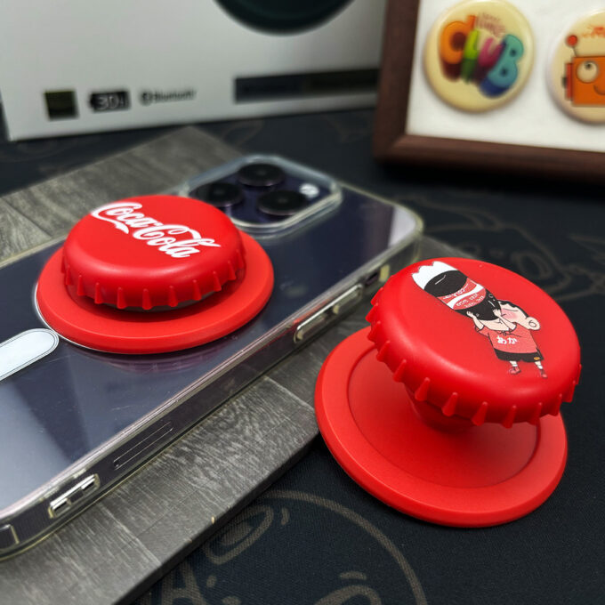 Two Coca-Cola bottle cap grips snapped onto a silver phone, with a separate red cap placed nearby, demonstrating their foldable functionality.