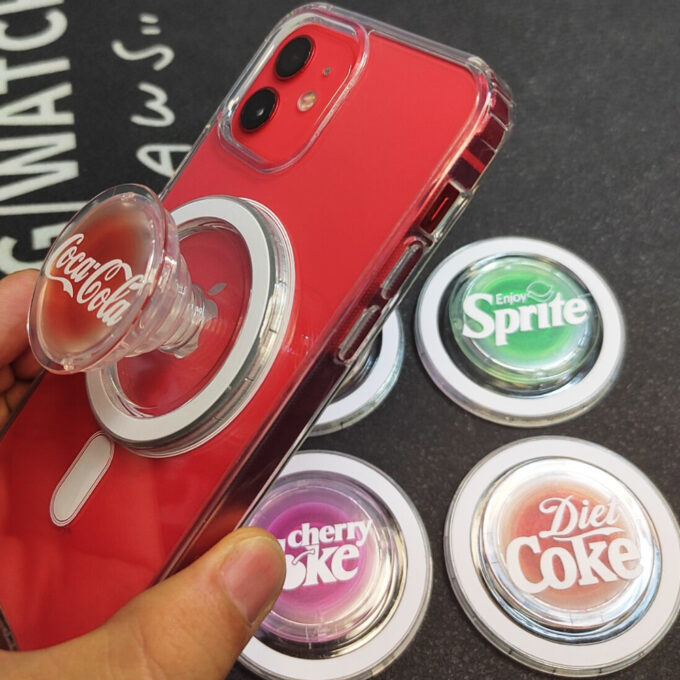 A red iPhone with a semi-translucent Coca-Cola MagSafe grip, surrounded by Sprite, Cherry Coke, and other branded grips.