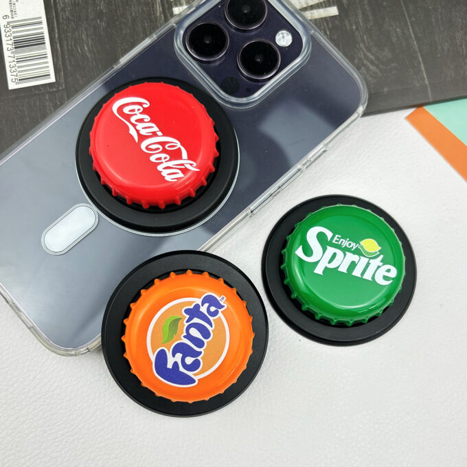 A gray iPhone with a Coca-Cola bottle cap grip attached, surrounded by additional caps in Sprite and Fanta designs.