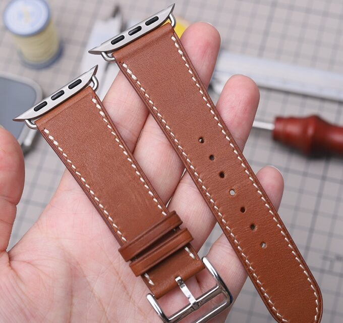 A brown French Swift Leather Apple Watch band with contrasting stitching, held up to display its smooth finish and stainless steel hardware.