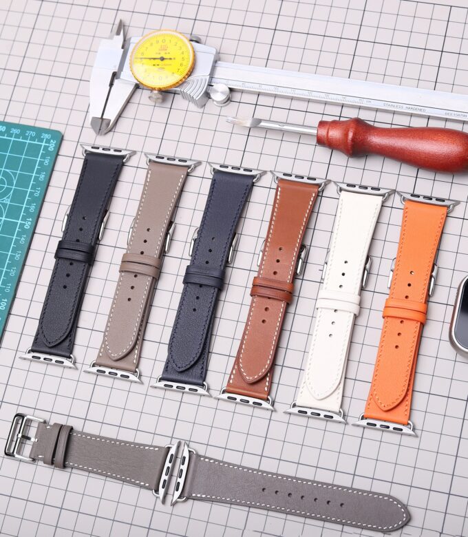 A selection of multiple Apple Watch bands in various colors (gray, brown, white, navy, orange), laid out on a grid mat alongside leathercraft tools.