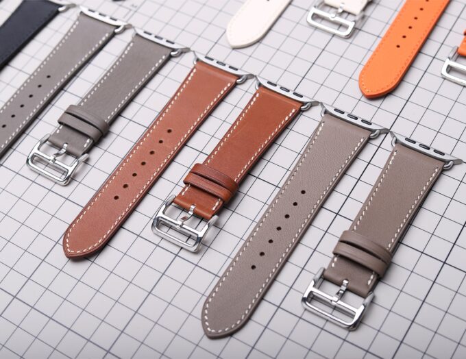 Several earth-toned Apple Watch bands lined up in a row, highlighting the genuine French leather and stainless steel hardware.