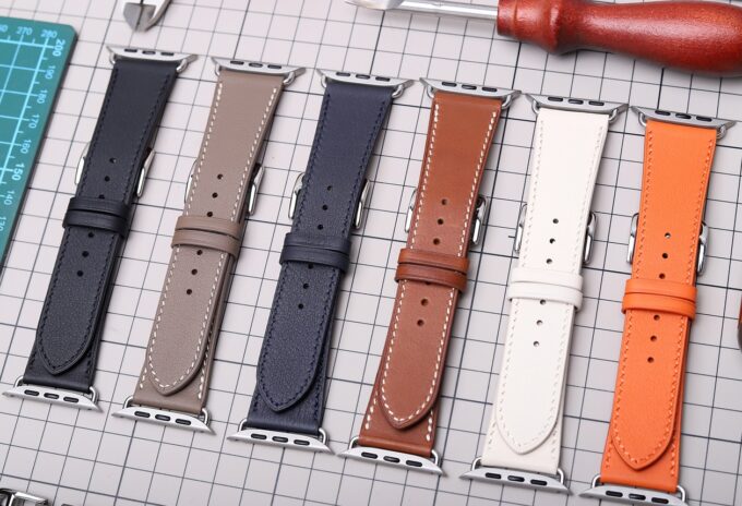 A mix of neutral (gray, brown) and vibrant (orange, white) leather Apple Watch bands displayed together, showcasing their flexibility and refined look.