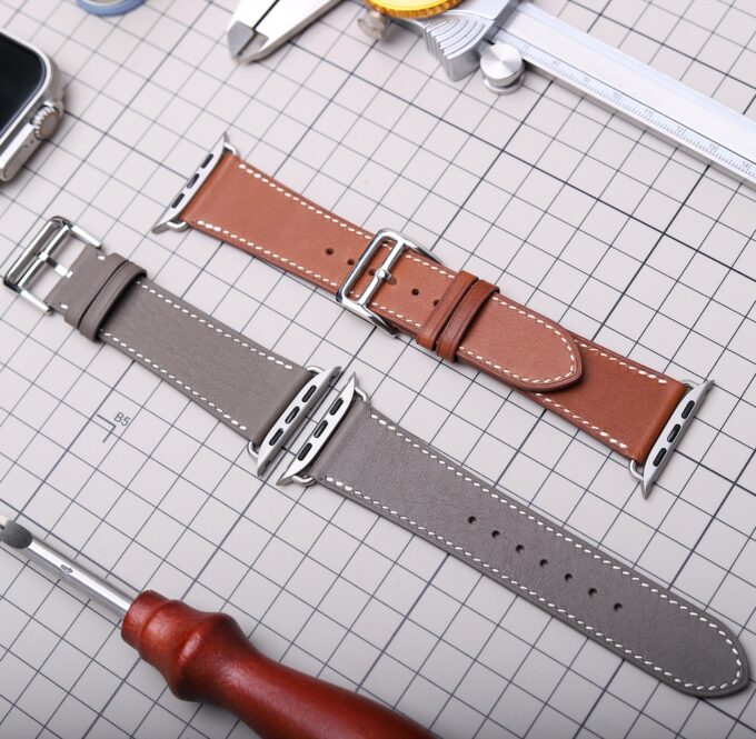 Two leather watch bands, each showing detailed stitching and a polished stainless steel buckle, laid out on a grid mat for a closer look.