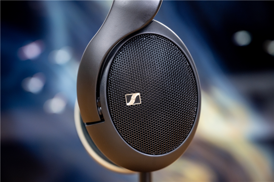 Close-up of the Sennheiser HD 505 open-back headphone's ear cup, showcasing the minimalist black design with the Sennheiser logo and subtle gold accents on the side, resting on a stand against a blurred background.